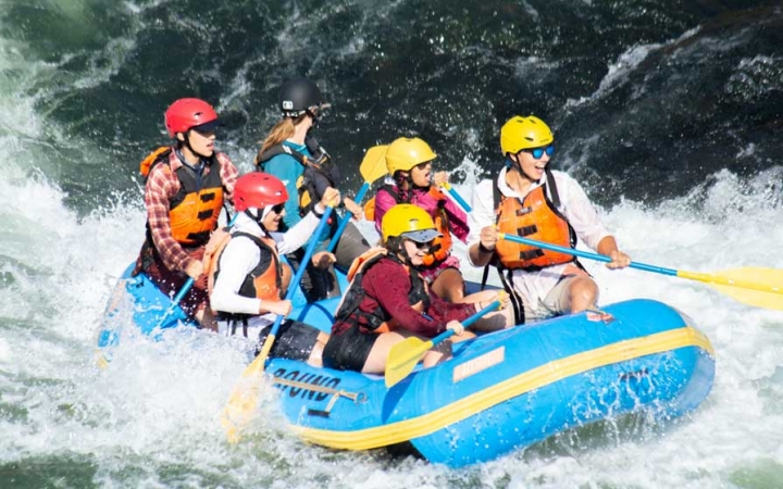 a group of outward bound students wearing helmets and life jackets paddle a raft through whitewater